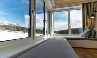 A modern living space with large windows that provide a view of a snow-covered landscape. Outside, mountains and a clear sky can be seen.