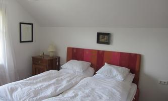 A cozy bedroom with a double bed and white bed covers. Pictures hang on the wall, and there is a nightstand with a lamp.