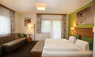 A modern hotel room with two beds, a sofa, and a large window. The walls are in soft colors and there are decorative elements like pictures and plants.