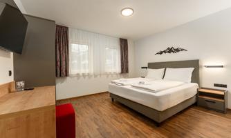 A modern hotel room with a large bed and light wood flooring. The room features a window with curtains and simple furnishings.