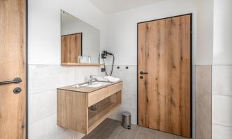 A modern bathroom with a wooden vanity and mirror. The walls are light and the door is made of wood.