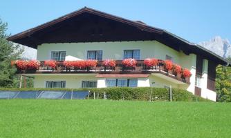 A traditional Alpine house with a balcony adorned with red geraniums. In the foreground, there is a green meadow and in the background, mountains are visible.