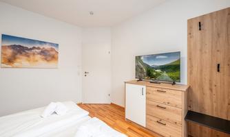 A bright bedroom with wooden flooring and modern furniture. A television sits on a dresser, while a painting hangs on the wall.