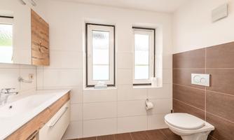 A modern bathroom with white tiles and wooden cabinets. Two windows provide plenty of light in the room.