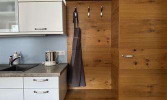 A modern kitchen with light cabinets and a wooden shelf. Utensils and an apron hang on the wall.