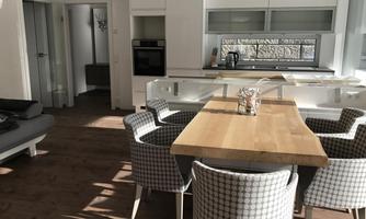 A modern kitchen with a large wooden table and upholstered chairs. There is plenty of natural light and a cozy atmosphere.