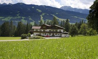 A picturesque landscape with a house, surrounded by green meadows and trees. In the background, tall mountains can be seen under a partly cloudy sky.