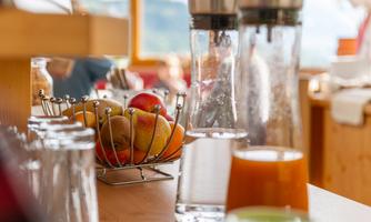 A table with glasses and water pitchers, surrounded by fresh fruit in a bowl. In the background, light wood and natural materials are visible.
