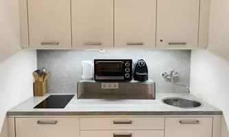 A modern kitchenette with white cabinets, a microwave, and a coffee machine. The room is bright and tidy, with a sink and a stovetop.