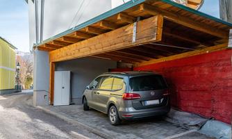 A gray car is parked under a wooden carport. In the background, a red wall and a tool shed are visible.