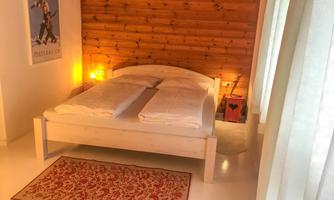 A bright bedroom with a double bed and a wooden wall. The room is cozily furnished and has a rug and curtains.