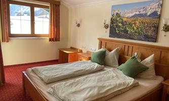 A cozy bedroom with two single beds, green pillows, and wooden furniture. A large window offers a view of the mountains.
