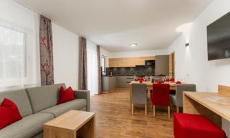 A modern living room with a grey sofa and red cushions. On one side, there is a dining area with a table and colored chairs.