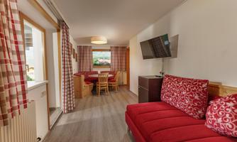 A cozy living space with red sofa cushions and a television. There is a dining area with a wooden table and chairs, as well as large windows for daylight.