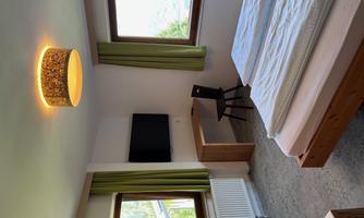 A modern bedroom with a double bed, a desk, and a television. The large windows offer a view of the outdoors and let in plenty of light.