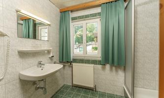 A simple bathroom with a shower, a sink, and a window. The walls are tiled, and the curtains are green.