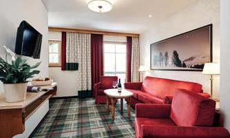 A cozy living space with red sofas and a round table. A television hangs on the wall, and the windows have beautiful curtains.