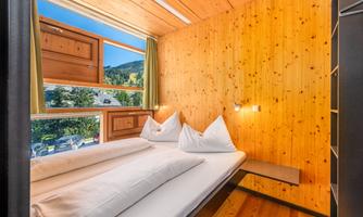 A bright bedroom with wooden paneling and large windows that offer a view of the mountains. The beds are simple, with white pillows.