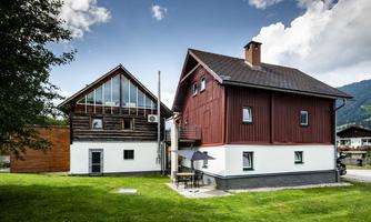 A modern house with a red facade and an adjacent wooden building. The sky is clear and the surroundings are green.
