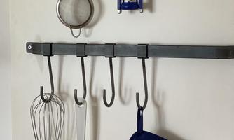 A wall with kitchen utensils hanging on a metal shelf. Below, a whisk, a spatula, and a blue pot holder can be seen.