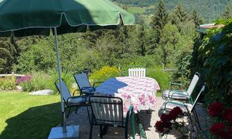 A beautiful garden with a table and chairs under a sun umbrella. In the background, there are green mountains and a blue sky.