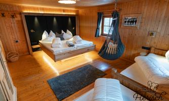 A cozy wooden room with two beds and a large double bed in the center. In the corner, there is a hanging swing, and the light is warm and inviting.