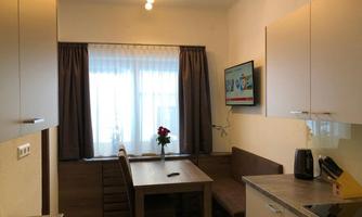 A cozy kitchen with a dining table and chairs. A TV on the wall and a window with curtains create a friendly atmosphere.