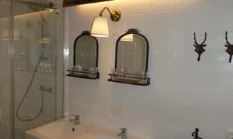 A modern bathroom with two sinks and two mirrors. The shower is separated by a glass wall, and decorative elements hang on the wall.