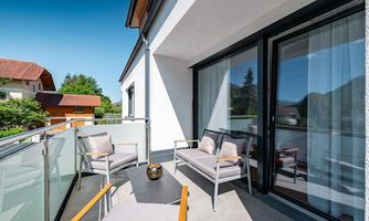 A modern balcony with two comfortable chairs and a low table. In the background, green trees and a sunny view can be seen.