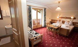 A cozy hotel room with red patterned carpet, a double bed, and a small table. On the left side, there is a bathroom with a toilet and sink.