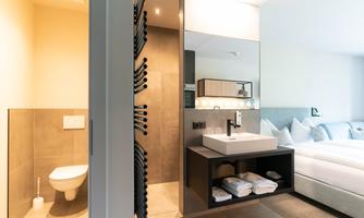 A modern room with a bathroom and a sink. The design is bright and minimalist, featuring a comfortable bed arrangement.