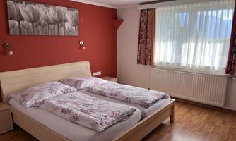 A cozy bedroom with a double bed and wooden flooring. The walls are painted in warm colors and a large window lets in plenty of daylight.