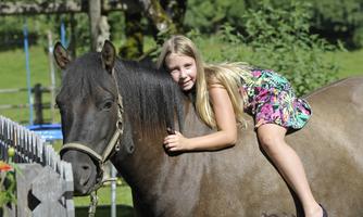 A girl is sitting on a horse and smiling. In the background, a green meadow and trees can be seen.