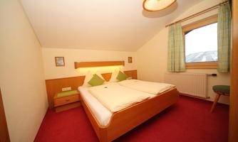 A cozy bedroom with two single beds and a red carpet. The room has a window with a view and bright curtains.