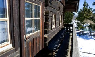 A traditional wooden house with a balcony, surrounded by snow and fir trees. The windows let in a lot of light and create a cozy atmosphere.