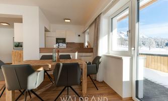 A modern living room with a dining table and elegant chairs. Large windows offer a view of the snowy landscape outside.