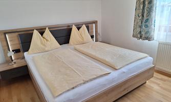 A cozy bedroom with a large double bed and two pillows. The room has bright walls and window decor with floral patterns.