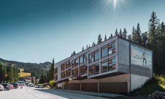 A modern building in the mountains with many windows. Surrounding it are trees and a parking lot.