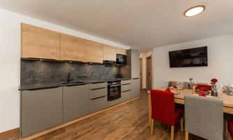 Modern kitchen with wooden and grey cabinets. A dining area with red and grey chairs, as well as a television, is also visible.