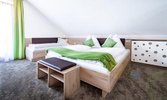 A bright and modern bedroom with a large bed and an additional single bed. The furnishings are made of wood with green accents and large windows for plenty of natural light.