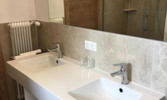 A modern bathroom with a large double sink and two washbasins. In the background, there is a shower and a towel holder.