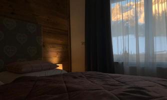 A cozy bedroom with a large window and a view of the mountains. Soft bed linens and a warm atmosphere create an inviting mood.