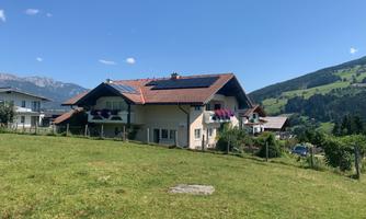 A beautiful house with a red roof in a green landscape. In the background, mountains and a clear blue sky can be seen.
