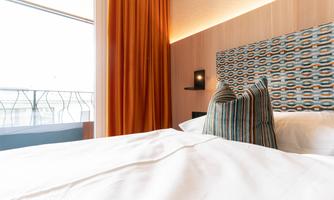 A modern bedroom with a large bed and colorful pillows. The curtains are orange and a lot of light comes through the window.