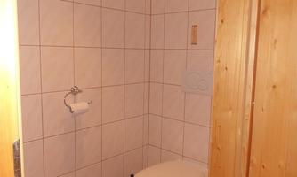 A small bathroom with a toilet and a gray bath mat. The walls are tiled with light tiles.