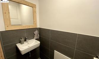A modern bathroom with a white sink and a wooden frame mirror. The walls are painted in neutral shades of gray.