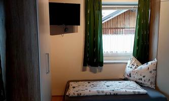 A simple bedroom with two beds, a television, and a window. The walls are light, and the curtains are green.