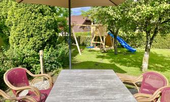 A cozy garden with a table and chairs under a sunshade. In the background, a slide and a playground can be seen.