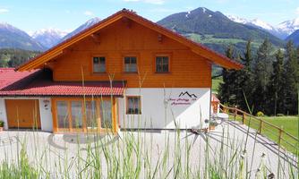 A cozy house in the mountains with wooden siding and red roof tiles. In the background, green meadows and snow-covered peaks are visible.