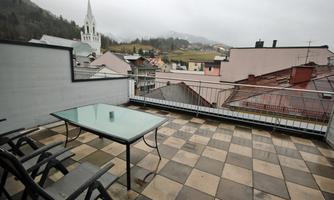A balcony with a glass table and chairs that offers a view of the rooftops of a city. In the background, mountains and a church can be seen under a gray sky.
